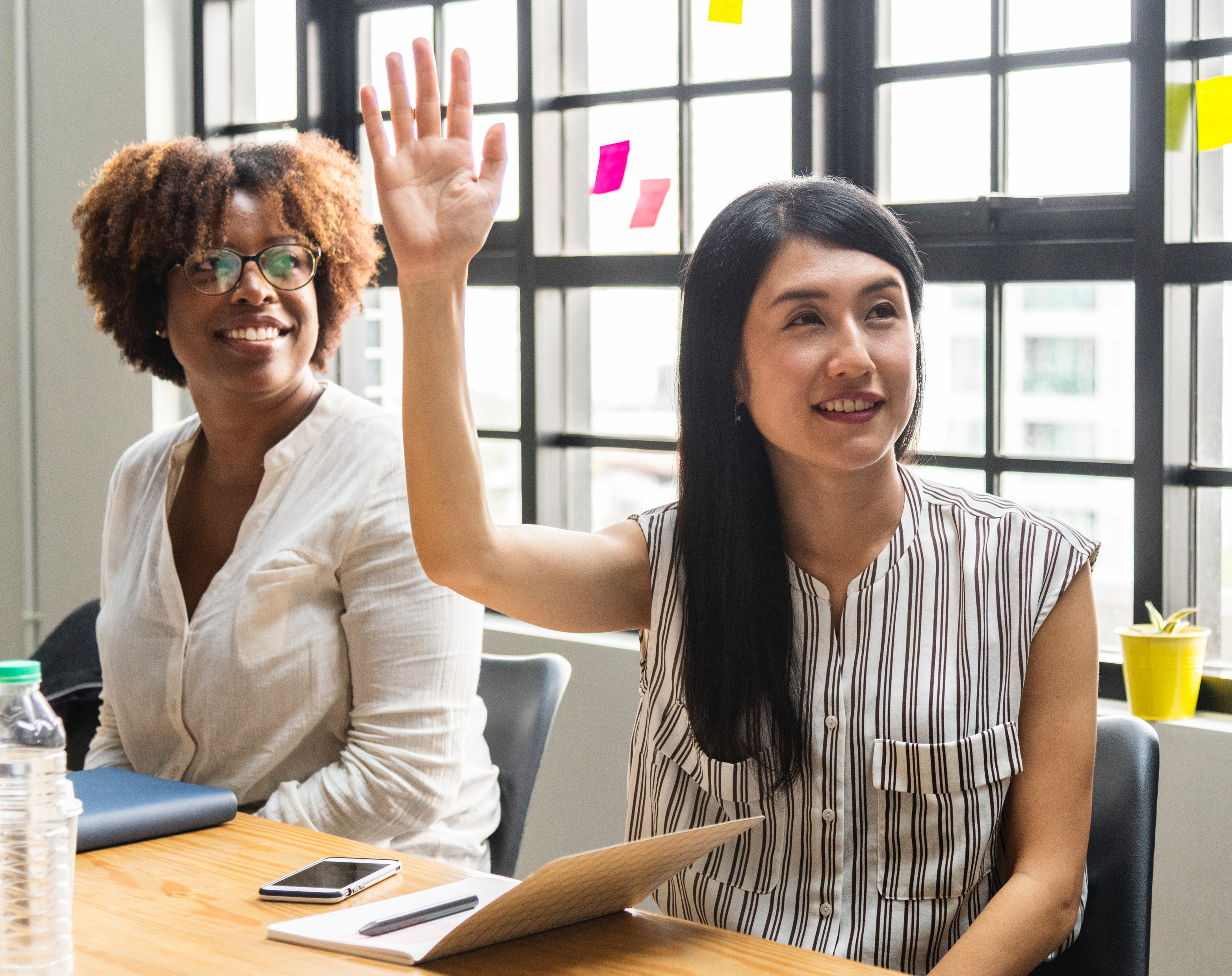 Figure 11: The image shows two people sitting at a table. One of the people has their hand raised to ask a question. The image is by rawpixel.