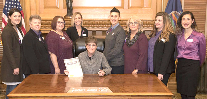 The image shows eight KCSDV staff members and coalition member program staff with Kansas Governor Laura Kelly at the 2019 Domestic Violence Awareness Month Proclamation Signing Ceremony in Topeka. The image is by the Kansas Governor’s Office and edited by KCSDV.