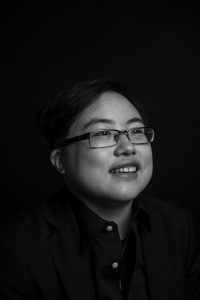 Photo: Black and white image of Lydia X. Z. Brown, a young East Asian person with glasses, smiling and laughing, looking slightly away from the camera. Photo by Colin Pieters.